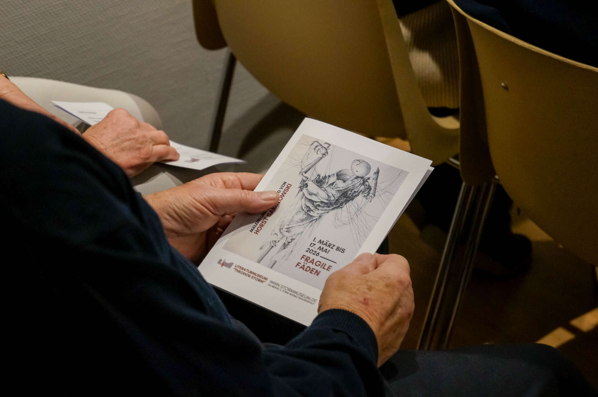 Der Flyer zur Ausstellung wird von einer Person in den Händen gehalten