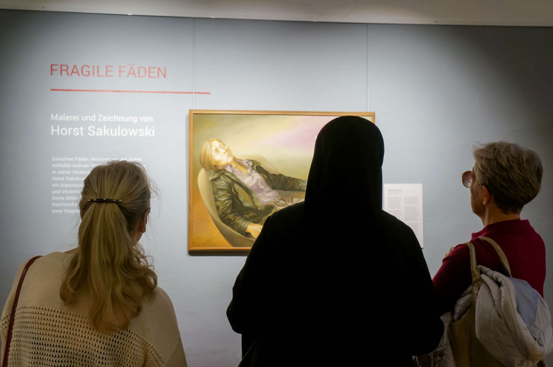 Drei Personen betrachten ein Bild der Ausstellung. An der Wand ist Text. Er zeigt den Ausstellungstitel "Fragile Fäden".