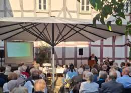 Sitzendes Publikum von hinten fotografiert, die auf eine Frau am Rednerpult schauen.